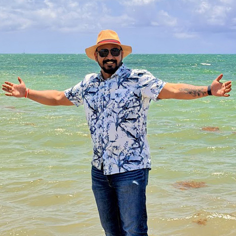 Avijit Roy smiling with arms outstretched on a beach in Key West, wearing a straw fedora hat, blue floral shirt, and sunglasses with the ocean in the background.